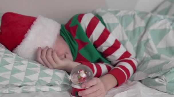 Un garçon dans une casquette du Père Noël se trouve au lit le jour du Nouvel An avec une boule de verre de neige jouet — Video