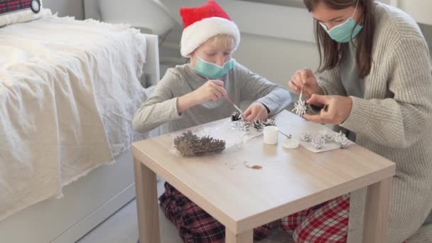Mother and son decorate spruce cones with white acrylic paint in protective medical masks. — Stock Video