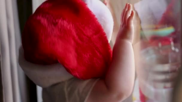 Um close-up de um garoto em um boné Papai Noel olha pela janela — Vídeo de Stock