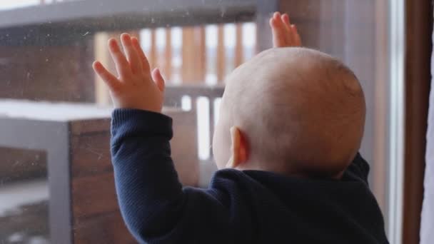 En söt liten pojke tittar ut genom fönstret och träffar glaset med händerna — Stockvideo