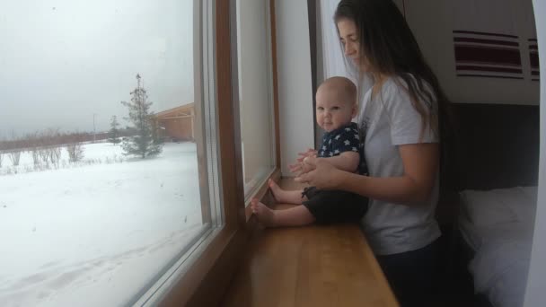 Uma jovem mãe beija seu filho e olha pela janela para uma rua coberta de neve durante — Vídeo de Stock
