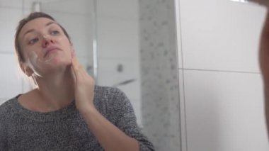 Bir kadın sabah banyoda aynanın önünde yüzüne nemlendirici sürüyor.