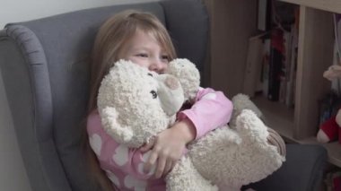 Sevimli küçük bir kız sevgili oyuncak ayısını evinde bir sandalyede kucaklıyor.