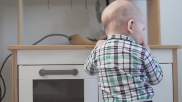 Un niño pequeño en una camisa se para con el apoyo en la cocina del juguete de los niños — Vídeos de Stock