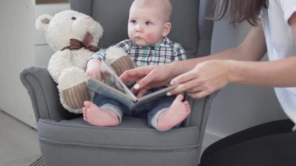 Mama pokazuje książkę z obrazkami małemu dzieciakowi w domu na fotelu dla dzieci — Wideo stockowe