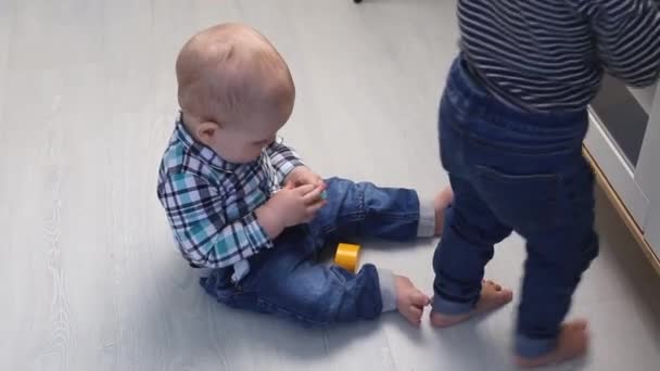 Un niño pequeño en una camisa se sienta en el suelo de la casa y lame un juguete pequeño — Vídeo de stock