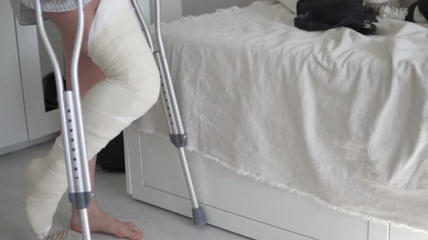 Une femme avec du plâtre sur la jambe droite sur des béquilles pour s'asseoir sur le lit — Video