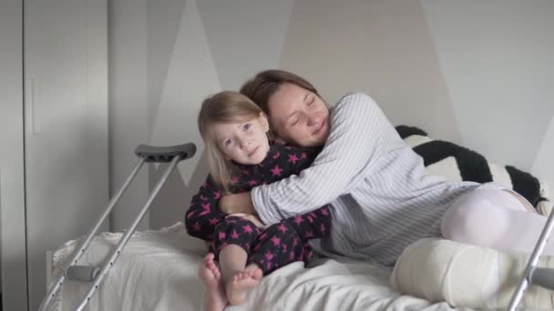 Мать со сломанной ногой в гипсе обнимает свою маленькую дочь дома в постели — стоковое видео