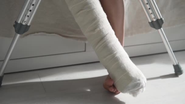 A woman in plaster on her leg sits on the bed and lifts her legs up to heal — Stock Video