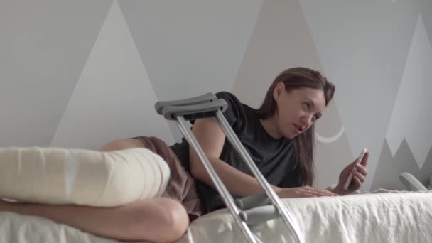 O femeie frumoasă cu piciorul bandajat se află pe pat și vorbește la telefon prin intermediul unui link video în spital. — Videoclip de stoc