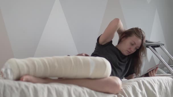 Une belle femme avec un énorme plâtre sur la jambe se trouve sur le canapé et utilise une tablette avec Internet — Video