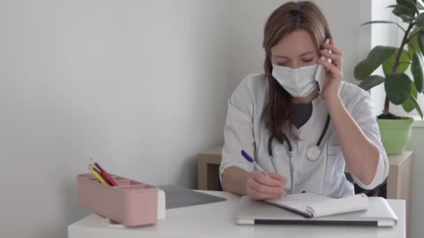 A masked doctor talks on the phone and invites the client to enter the office — Wideo stockowe