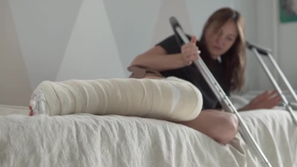 Una mujer con una pierna rota pone muletas y se va a la cama — Vídeo de stock
