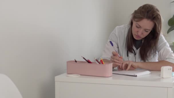 Uma bela médica vestida com um manto branco faz anotações em uma mesa no estudo — Vídeo de Stock