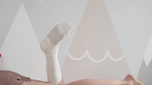 Uma mulher com gesso na perna repousa na cama e levanta a perna — Vídeo de Stock