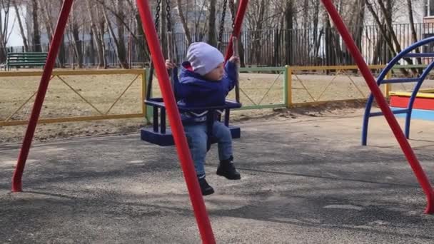 Uma criança pequena bonito senta-se em um balanço no playground na primavera — Vídeo de Stock