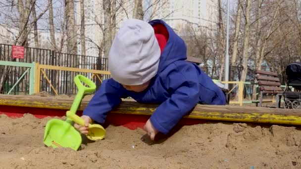 Egy kisfiú csöpög a homokban egy gyerekkel lapátol egy homokozóban tavasszal. — Stock videók