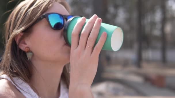Een vrouw met een zonnebril drinkt in het voorjaar koffie uit een plastic glas op straat — Stockvideo