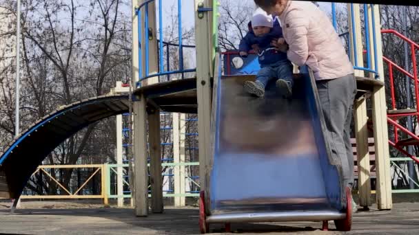 Una madre cavalca un bambino da una collina in un parco giochi per bambini in primavera — Video Stock