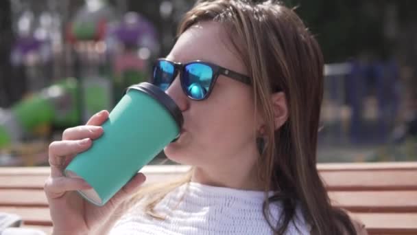 Uma mulher com óculos de sol bebe café na rua ao lado do playground — Vídeo de Stock