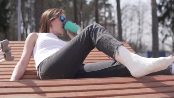 En vacker kvinna med gips på benet dricker kaffe i parken på våren — Stockvideo