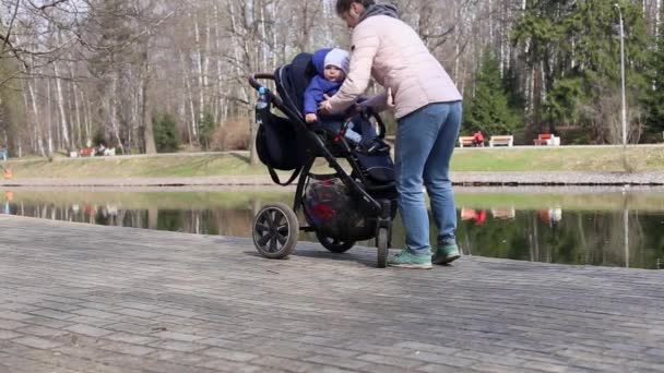 Egy fiatal anya betesz egy kisgyereket egy babakocsiba egy séta során a parkban tavasszal. — Stock videók