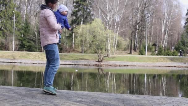 Μια μητέρα κουβαλάει ένα μικρό παιδί στην αγκαλιά της κατά τη διάρκεια μιας βόλτας στο πάρκο την άνοιξη — Αρχείο Βίντεο