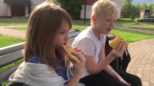 Az éhes gyerekek gyorskaját esznek a parkban egy padon. — Stock videók