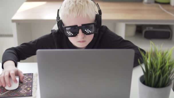 Ein Junge mit lustiger Brille und Kopfhörer spielt zu Hause an seinem Laptop ein Computerspiel — Stockvideo