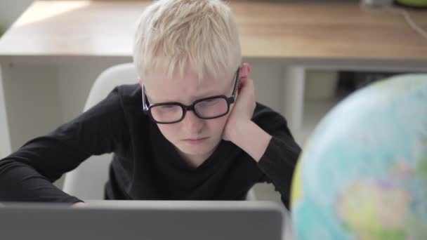 Un niño con gafas se sienta en un escritorio en la escuela y entrena en una computadora — Vídeos de Stock
