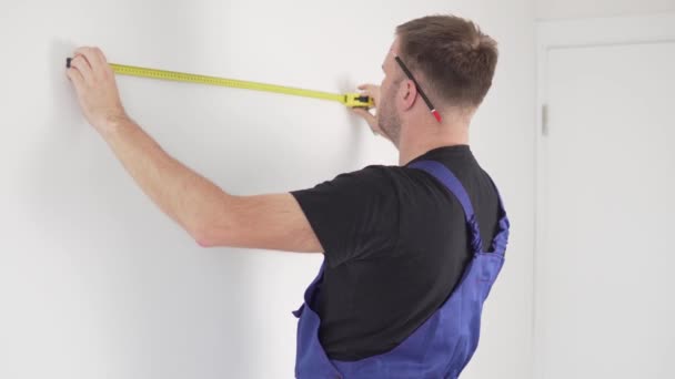 Een mannelijke arbeider meet de stip op de muur met een centimeter roulette en potlood — Stockvideo