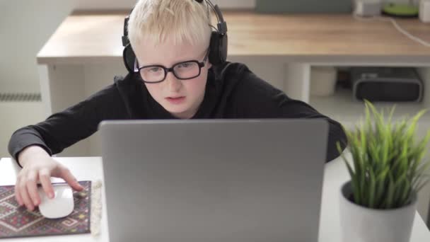 Ein Junge mit Brille und Kopfhörer spielt zu Hause am Tisch mit einem Laptop — Stockvideo