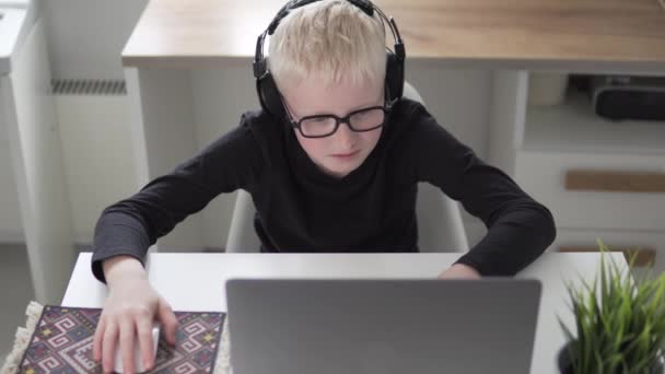 Ein glückliches Kind mit Kopfhörern und Brille spielt von zu Hause aus ein Computerspiel im Netz — Stockvideo