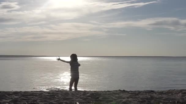 Une petite fille joue dans l'eau au coucher du soleil sur la plage au bord du lac — Video