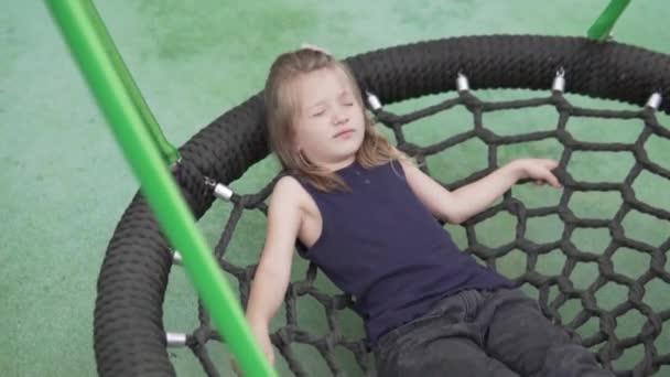 Una bella bambina si pente su un'altalena sotto forma di ragnatela nel parco giochi in estate. — Video Stock