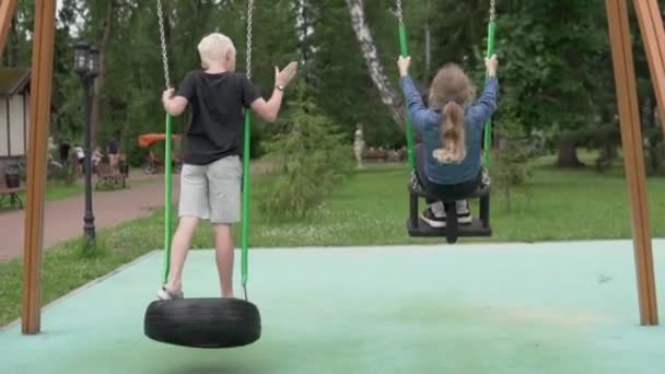 Дети школьников катаются на качелях в парке летом, вид сзади — стоковое видео