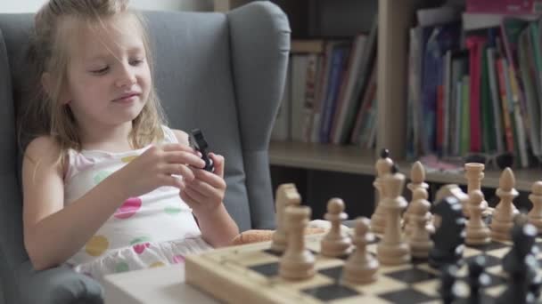 Ένα μικρό κορίτσι μασάει ένα πιόνι στο σκάκι κατά τη διάρκεια μιας τάξης. — Αρχείο Βίντεο