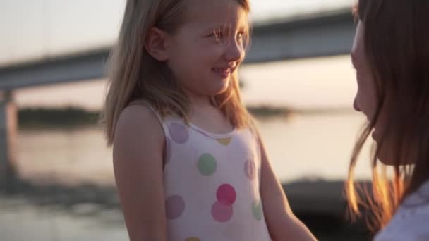 Talking to a mother with a little girl at sunset by the river in the summer, the daughter hugs her mother — Stock Video