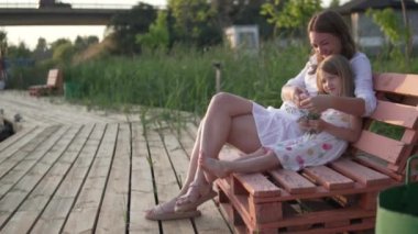 Bir anne ve küçük bir kız gün batımında tahta bir bankta kucaklaşırlar.