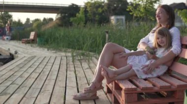 Güzel anne ve kız yazın beyaz elbiselerle parkta bir bankta oturur ve gün batımını beklerler.