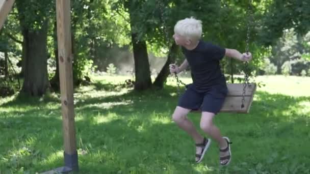 Een albino jongen rijdt een schommel in de zomer in een schaduwrijk park — Stockvideo