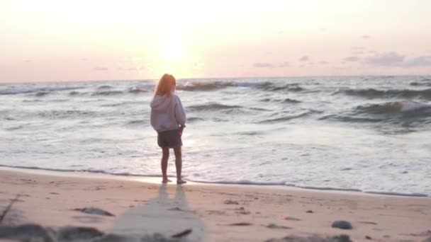 Una niña está de pie con su espalda al mar y observa la puesta de sol en una playa de arena desierta — Vídeos de Stock