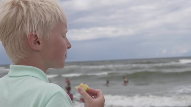 Un garçon blond mange une pomme sur la plage au bord de la mer — Video