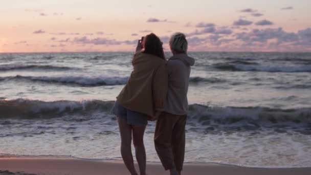 Duas amigas tiram uma foto na praia ao pôr-do-sol, uma vista pelas costas — Vídeo de Stock