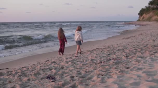 Δύο χαριτωμένα μικρά περπατούν κατά μήκος της παραλίας κατά μήκος της θάλασσας το ηλιοβασίλεμα. — Αρχείο Βίντεο