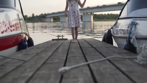 Una niña corre a lo largo de un puente de madera entre yates en la noche al atardecer — Vídeos de Stock