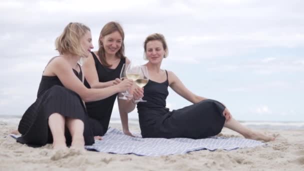 Três mulheres bonitas em vestidos pretos bebem vinho na praia pelo mar de óculos — Vídeo de Stock