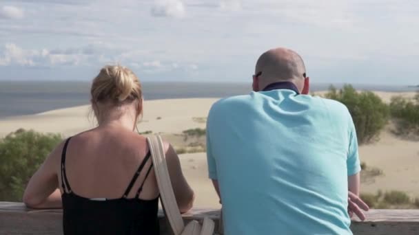 Vista desde atrás, un hombre y una mujer mirando el mar y las dunas de arena desde la plataforma de observación de vacaciones — Vídeo de stock