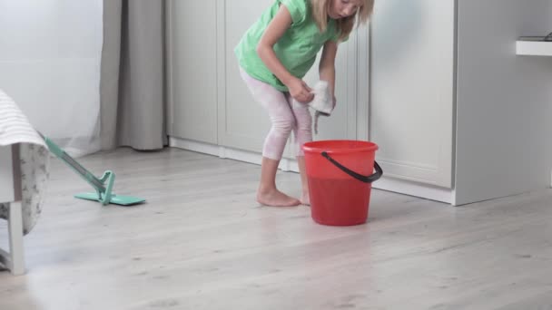 Ein kleines Mädchen wäscht den Fußboden des Hauses mit einem Sextuch, das Baby wäscht einen schmutzigen Lappen in einem Eimer Wasser — Stockvideo