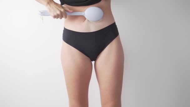 Parte das pernas femininas e barriga, ela faz massagem seca anti-celulite em seu corpo com escova em um fundo branco da casa — Vídeo de Stock